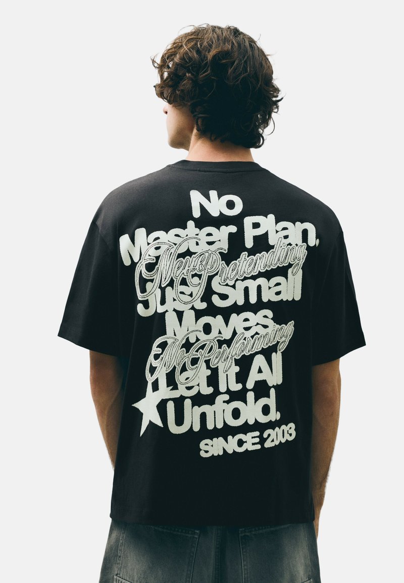 Person with curly hair stands facing away wearing black t-shirt with large white mixed-font text on the back.