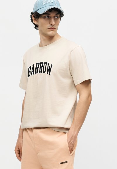 Νέος άνδρας που φοράει μπεζ μπλουζάκι Barrow και ροδακινί σορτς με το λογότυπο Barrow, ανοιχτό μπλε τζιν καπέλο, στέκεται με το ένα χέρι στην τσέπη.