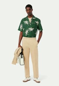 Camisa verde de manga curta com estampa abstracta branca, calças bege e ténis. Inclui uma jaqueta leve e uma bolsa em preto e branco.