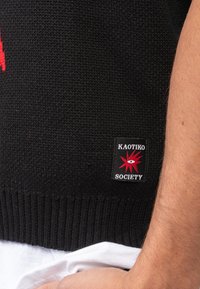 Μαύρο πλεκτό πουλόβερ με ριγωτό τελείωμα και κόκκινο λογότυπο που γράφει "KAOTIKO SOCIETY". Το υλικό έχει υφή με χαλαρή ύφανση.