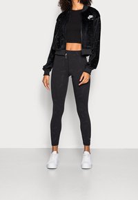 Kvinna iklädd svart Nike Air kort jacka, svart crop top, svarta leggings med subtila logotyper, och vita sneakers, stående mot vit vägg.