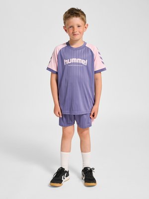 Ung dreng står med fronten mod kameraet iført lilla og pink sportstøj med "hummel" logo, hvide strømper og sorte sneakers på ensfarvet baggrund.