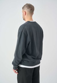 Black sweatshirt with a relaxed fit, ribbed cuffs, and hem. Layered over a white shirt, displayed from the back. Soft texture and casual design.