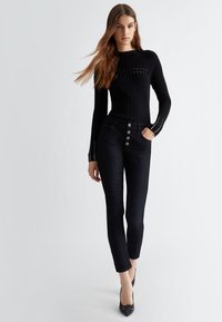 Maglietta nera a costine con maniche lunghe e dettagli a taglio, abbinata a pantaloni in denim scuri con chiusura a cinque bottoni e vestibilità sartoriale.