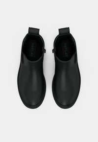 Para czarnych skórzanych botków za kostkę typu slip-on z uchwytami i subtelnym detalem w postaci zamka, widoczna z góry na białym tle.