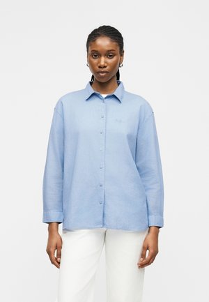 Γυναίκα με πλεξούδες, που φοράει ανοιχτό μπλε πουκάμισο με κουμπιά και λευκό παντελόνι, στέκεται μπροστά σε απλό φωτεινό φόντο.