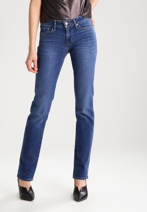 Person trägt mittel-hohe, schmal geschnittene blaue Jeans und schwarze spitz zulaufende Absätze und steht vor einem einfarbigen hellen Hintergrund.