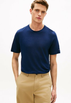 Giovane uomo che indossa una maglietta a maniche corte blu navy e pantaloni beige, in piedi con le mani nelle tasche contro uno sfondo semplice.