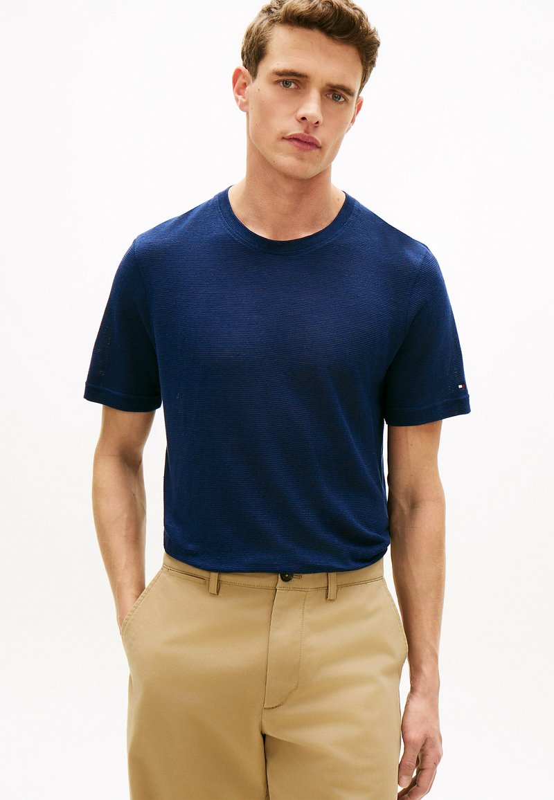 Jeune homme portant un pull à manches courtes bleu marine et un pantalon beige, debout les mains dans les poches devant un fond uni.