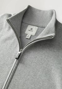 Grauer Kapuzenpullover mit Reißverschluss, ausgestattet mit einem gerippten Kragen und einem Reißverschluss. Mit einem Stoffetikett, das "NEXT" und die Größenangaben zeigt. Glatte Textur.
