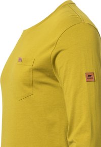 Gelbes Langarmshirt aus weichem Stoff mit einer Brusttasche und einem Lederlabel am Ärmel mit einem Bärenlogo.
