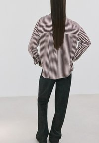 Persona con cabello largo y liso que lleva una camisa holgada a rayas burdeos y blancas, pantalones oscuros, de pie con las manos en las caderas y de espaldas.
