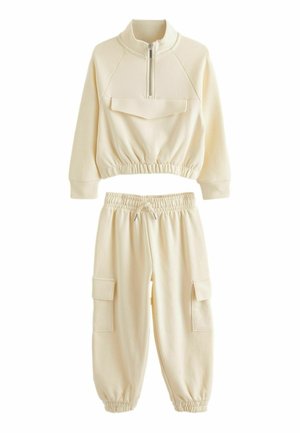 Kreminės spalvos deranti sportinė apranga, pagaminta iš minkštos medžiagos. Viršutinė dalis turi užtrauktuką ir priekinius kišenes; kelnės turi elastingus rankogalius ir cargo kišenes.