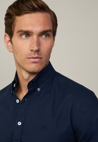 Navyblauw overhemd met kraag en knoopsluiting, voorzien van een klein geborduurd embleem op de borst, gladde stofstructuur en contrasterende knopen.