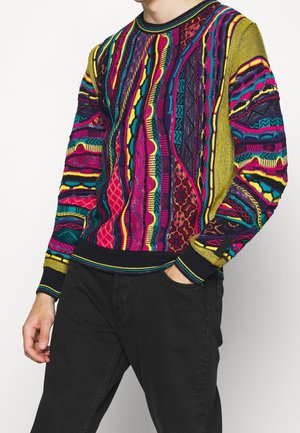 Homme portant un pull coloré à motifs avec des détails roses, jaunes, bleus et noirs et un pantalon noir, une main dans la poche.