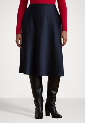 Femme portant une jupe midi noire associée à des bottes en cuir noires à talons et un haut rouge à manches longues, debout sur un fond uni.