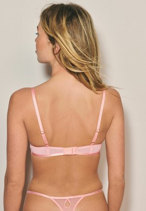 Woman with blonde hair wearing a light pink bra and matching thong, facing away against a plain beige background.