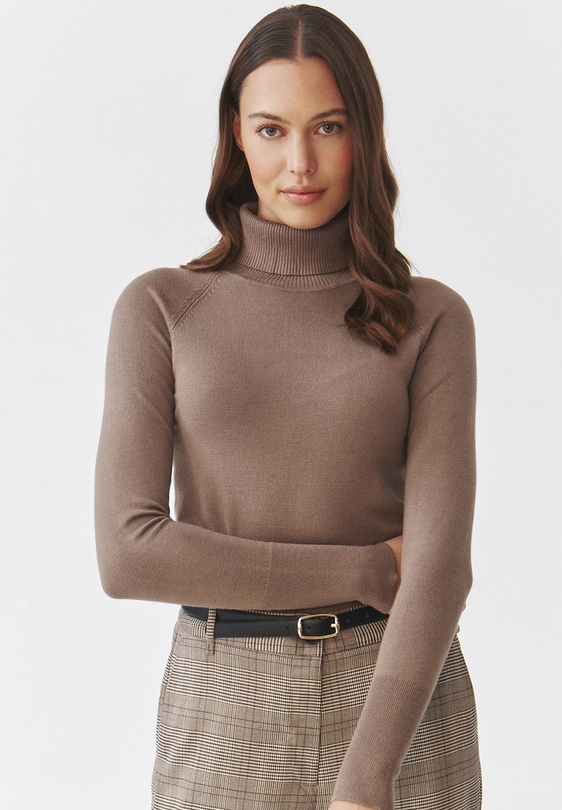 Brązowy sweter typu golf z długimi rękawami i prążkowanymi mankietami, w połączeniu z w kratkę spodniami oraz czarnym paskiem z złotą klamrą.