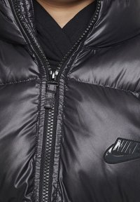 Chaqueta acolchada negra con cuello alto, que cuenta con un cierre de cremallera de largo completo y un logo de Nike brillante en el pecho. Material suave y brillante.