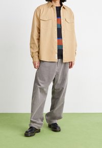 Vaaleanruskea napillinen paita, jossa rintataskut, päällä raidallinen musta, oranssi ja sininen T-paita. Harmaat corduroy-housut ja mustat kengät.