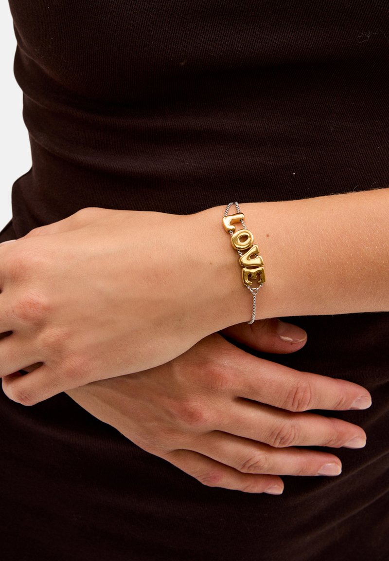 Bracciale tono oro con lettere in blocco che formano la parola "AMORE", attaccato con una catena argentata, indossato su un polso contro uno sfondo di tessuto scuro.