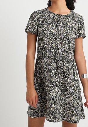 Femme portant une robe noire à manches courtes, longueur genou, avec un petit motif floral blanc et jaune, et un bracelet manchette argenté sur son bras droit.