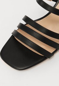 Sandal negru cu patru curele late, vârful pătrat și o branț de un bej deschis. Fabricat din piele ecologică netedă, cu cusături vizibile.