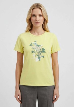 Donna bionda che indossa una t-shirt gialla a maniche corte con stampa floreale verde e bianca e pantaloni grigi, in piedi davanti a uno sfondo semplice.