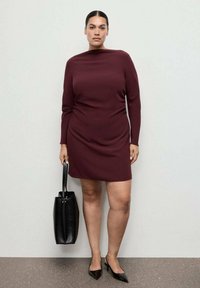 Vestido burdeos de manga larga con cuello redondo, silueta ajustada y detalle drapeado, acompañado de un bolso tote negro y zapatos de punta.