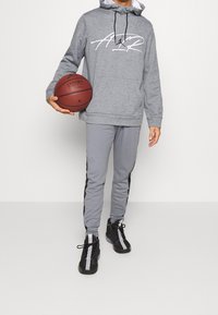 Felpa grigia con logo bianco, pantaloni da jogging grigi con strisce nere laterali e sneakers nere. Tiene in mano un pallone da basket. Outfit sportivo e casual.