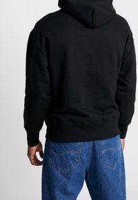 Felpa nera con un ampio cappuccio, polsini a costine e vita. La vista posteriore mostra una texture liscia. Abbinata a jeans di denim blu con tasche posteriori.