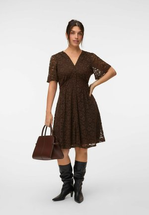 Femme portant une robe en dentelle marron à manches courtes, tenant un sac à main marron foncé et portant des bottes noires souples montant jusqu'aux genoux.