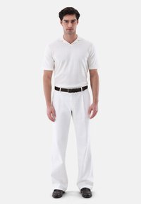 Weißes kurzärmliges Poloshirt mit V-Ausschnitt, kombiniert mit weit geschnittenen weißen Hosen und einem schwarzen Gürtel. Dunkle Schuhe runden das Outfit ab.