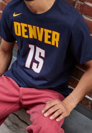 Mężczyzna ubrany w granatowy jersey Denver z numerem 15 i czerwone spodnie, siedzący oparty o ceglany mur z srebrną bransoletką na nadgarstku.