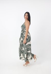 Maxi abito di un leggero colore beige con una stampa di foglie verde scuro. Presenta spalline sottili, vita arricciata e orlo fluente. Indossato con sandali bianchi con tacco.