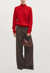 Rode geribbelde coltrui met schouderknopen, gecombineerd met donkerbruine wijde broek; model houdt een bruin, gebogen handtas vast.