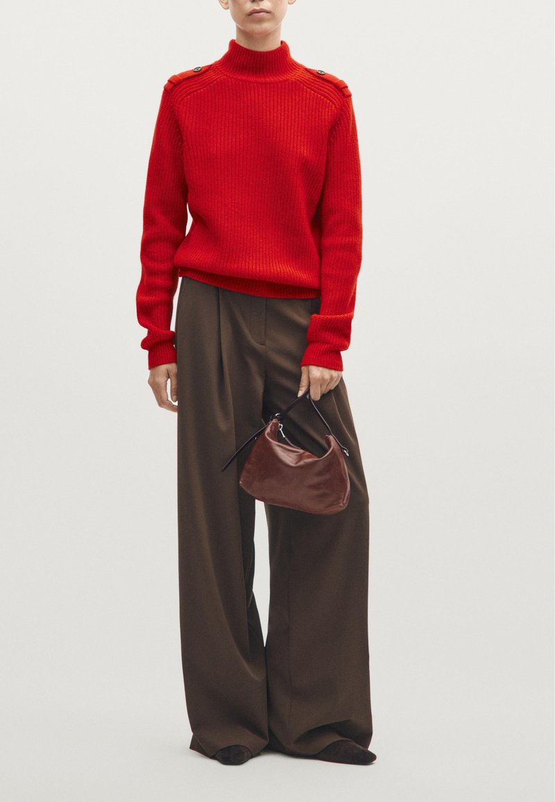 Maglione rosso a coste con collo alto e bottoni sulle spalle, abbinato a pantaloni larghi marroni scuri; la modella tiene in mano una borsa a tracolla curva marrone.
