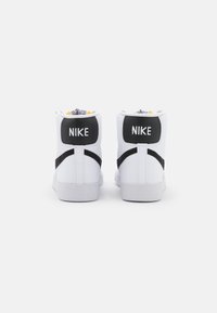 Høye hvite sneakers med svarte detaljer, utstyrt med doble svarte hæltabber merket "NIKE" og et teksturert svart stropp. Gummisåle, glatt overdel.