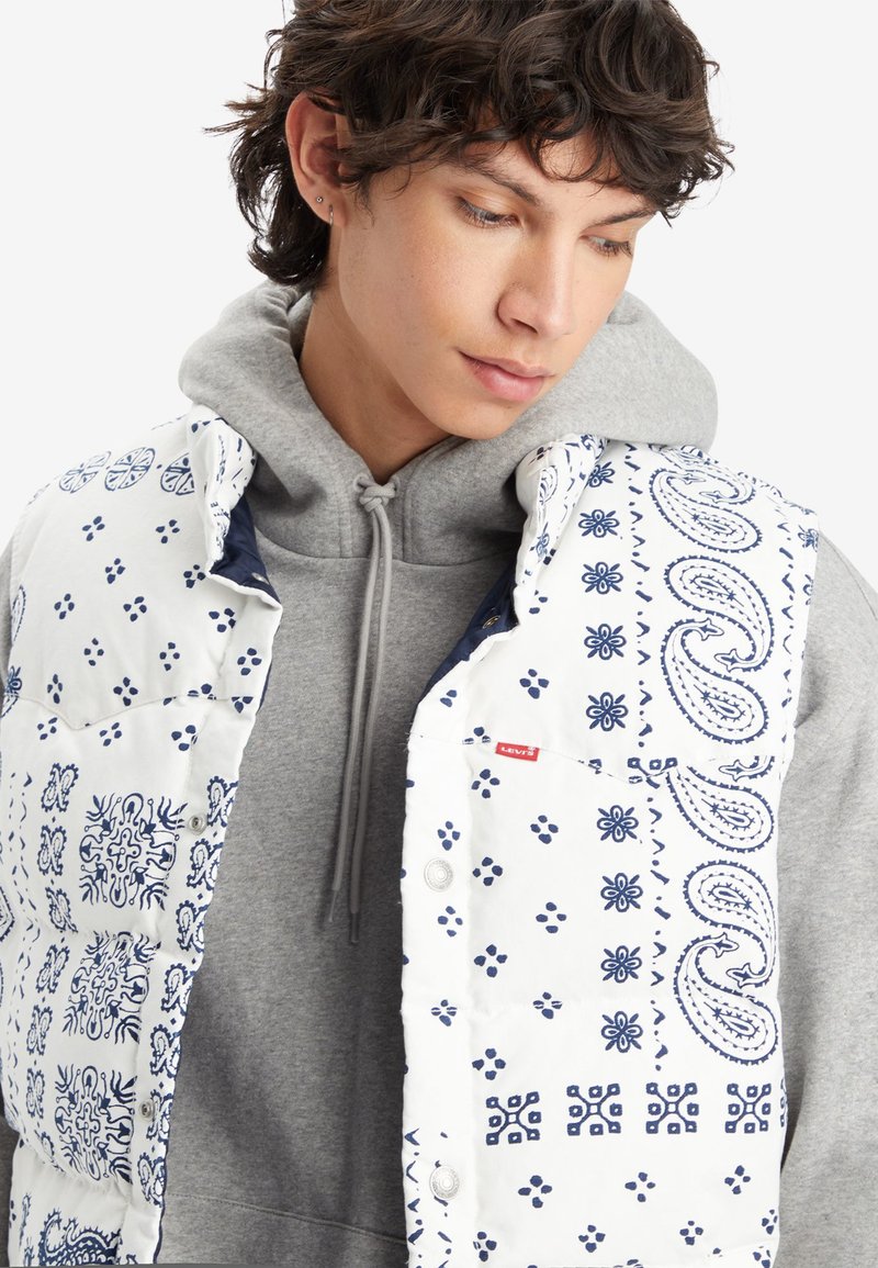 Chaleco acolchado blanco con patrones de paisley y flores azules, que cuenta con botones a presión y un logo de Levi's, superpuesto a una sudadera gris.