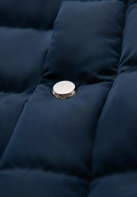 Mörkblå vadderad textil med en glänsande silverfärgad tryckknapp. Texturen verkar mjuk och slät, vilket framhäver det kviltade mönstret.