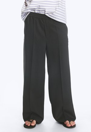 Pantalon large noir ample porté avec un haut rayé et des sandales tongs noires montrant les pieds nus.