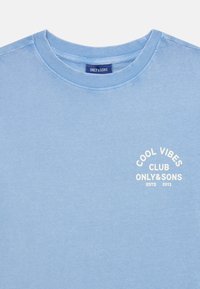 Světle modré bavlněné tričko s kulatým výstřihem, přední strana má bílé tištěné logo s nápisem "COOL VIBES CLUB ONLY&SONS ESTD 2013".