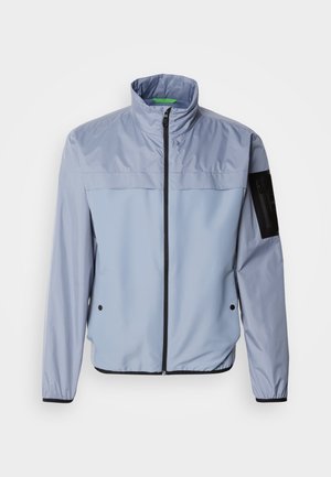 Veste bleu clair à manches longues avec un col haut, fermeture éclair sur le devant et accents noirs. Présente une poche sur la manche gauche et des poignets élastiques.