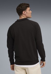 Pulover negru cu guler, având un design cu mâneci raglan și manșete cu dungi. Textură subtilă, completată de un maieu alb simplu.
