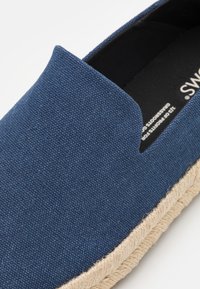 Scarpa slip-on in tela blu con suola in juta intrecciata. La parte superiore presenta un tessuto testurizzato e un'apertura laterale per la traspirabilità.
