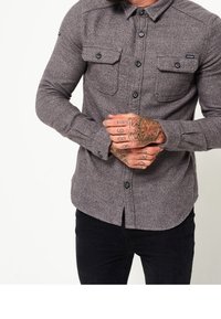 Camisa de franela gris con mangas largas, cierre delantero de botones, dos bolsillos en el pecho y botones negros. Presenta un collar clásico y un tejido texturizado.