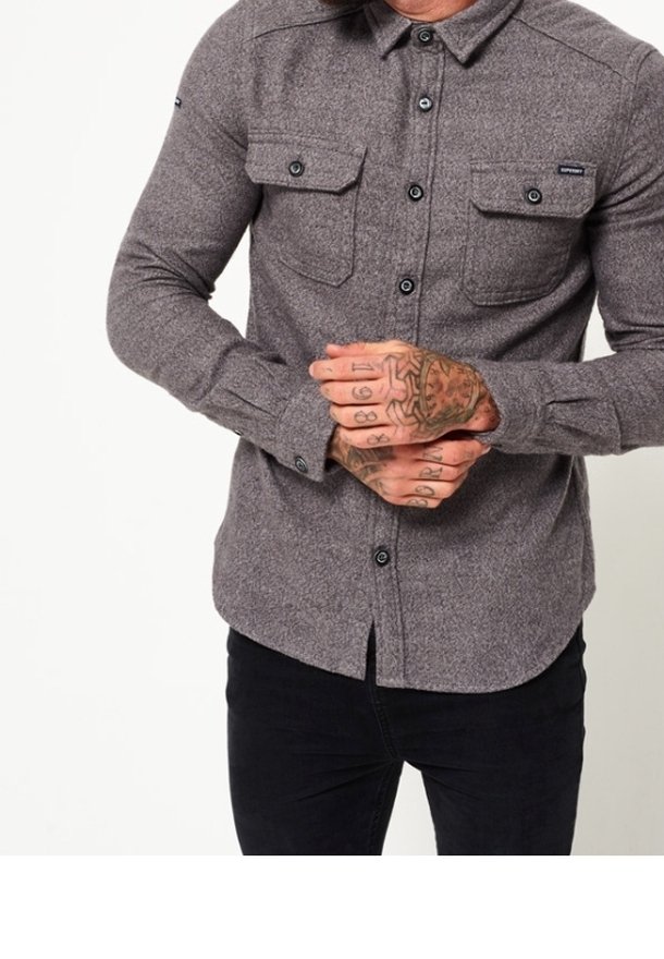 Camisa de franela gris con mangas largas, cierre delantero de botones, dos bolsillos en el pecho y botones negros. Presenta un collar clásico y un tejido texturizado.