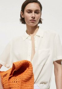 White, short-sleeve blouse with a button-down front and a chest pocket; paired with a textured orange crocheted bag.