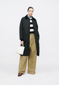 Czarny pikowany długi płaszcz z zatrzaskami na guzikach, na czarno-białym pasiastym swetrze, beżowe spodnie o szerokich nogawkach oraz mała biała torebka.