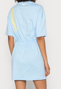 Light blue cotton dress with short sleeves, gathered waist, and yellow "Lacoste" logo printed vertically on the back. Smooth texture.